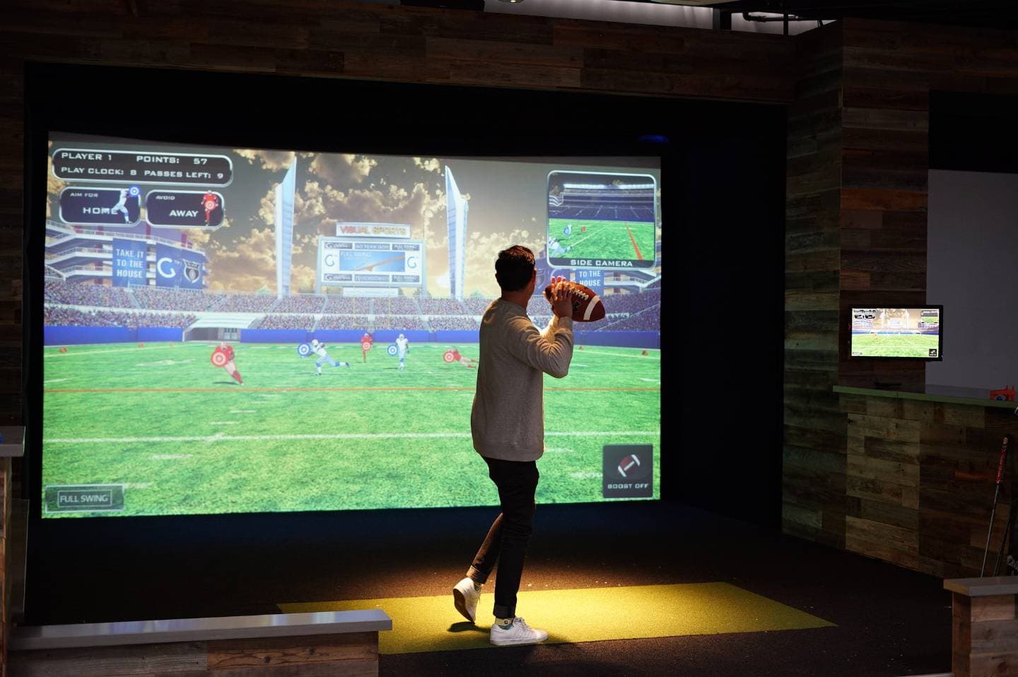 A player in a simulator bay throwing an American football at the screen, which shows a stadium pitch with receivers and target markers downfield.