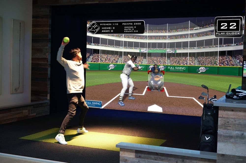 A player in a simulator bay pitching a ball at the screen, which shows a batter waiting at home plate in a floodlit stadium.